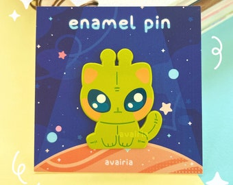 May include: A green alien cat enamel pin with large eyes and a pink nose. The pin is set against a dark blue background with stars and a planet. The text "enamel pin" is at the top of the card and the text "avairia" is at the bottom.