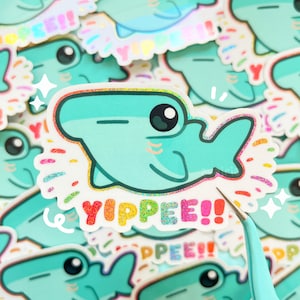 May include: A teal blue hammerhead shark sticker with a rainbow glitter finish. The sticker says "Yippee!" in rainbow glitter letters.