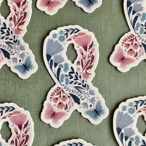 May include: A collection of stickers in a ribbon shape, adorned with floral and butterfly designs. The stickers feature a color palette of pink, blue, and burgundy, with a white background. The stickers are arranged on a green textured surface.