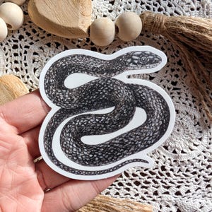 May include: A black and white sticker depicting a detailed illustration of a snake. The snake is coiled, with a textured pattern and a detailed head. The sticker is on a white background, held by a hand.