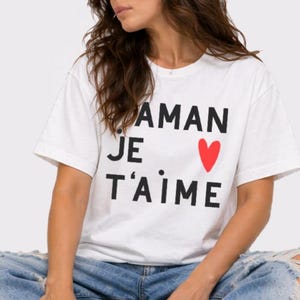 May include: White t-shirt with the French phrase "AMAN JE T'AIME" in black letters and a red heart. The model is wearing light blue ripped jeans. The shirt is a casual, everyday wear item.