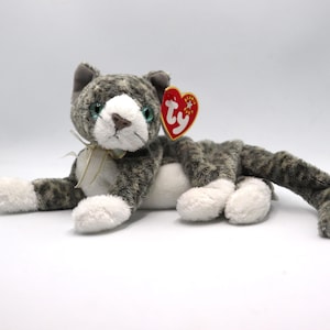 May include: A plush gray and white cat toy with a red heart-shaped tag. The cat has green eyes, a white muzzle, and a cream-colored ribbon tied around its neck. The cat's paws are white.