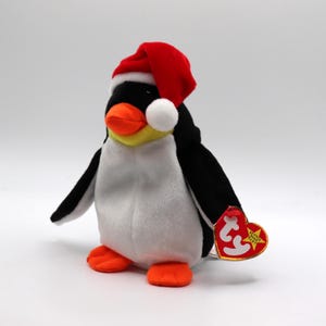 May include: A soft penguin toy wearing a red Santa hat, with a white pom-pom, and a heart-shaped tag. The penguin has a black body, white belly, orange feet and beak, and yellow accents. The tag reads "Beanie Baby".