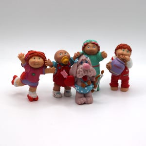 May include: A collection of Cabbage Patch Kids figurines. The figurines are in various poses and outfits, including a red-haired child with a cookie jar, a dog with a bow and arrow, and a baby with a pacifier.