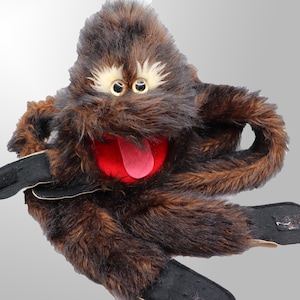 Op de afbeelding: Een bruine en zwarte monsterpop met een rode mond en roze tong. De pop heeft witte wenkbrauwen en twee gele ogen. De pop heeft zwarte handschoenen en zwarte voeten.