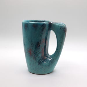 May include: A teal ceramic mug with a distinctive handle. The mug features a speckled texture with red and black detailing. The cylindrical mug is suitable for hot or cold drinks.