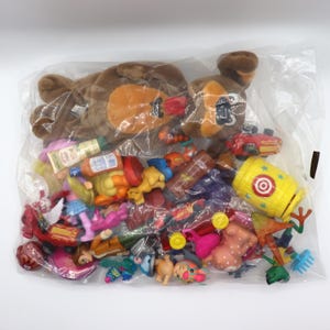 May include: A large clear plastic bag filled with a variety of assorted small plastic toys, including miniature figurines, toy vehicles, a yellow barrel with a target graphic, and a brown plush dog toy resting on top of the collection.