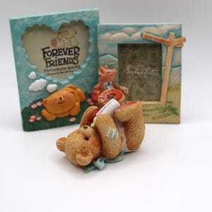 Può includere: Tre cornici decorative con statuette di orsi e maiali. Una cornice è azzurra con la scritta "Forever Friends". Un'altra cornice ha un cartello con la scritta "Over the Hills". Le statuette includono un orso che legge una pergamena.