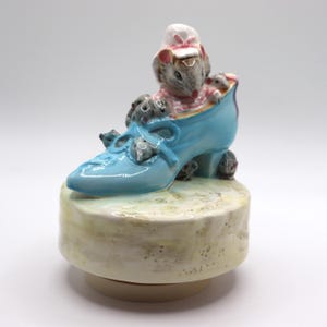May include: A ceramic figurine of a light blue shoe with a bow, filled with several gray mice. One mouse wears a pink and white outfit. The shoe sits on a round, off-white base with a yellow-green top.
