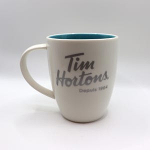 May include: A white ceramic mug with a curved handle, featuring the grey text Tim Hortons and the year 1964 printed on the front. The interior of the mug is a vibrant teal colour, providing a contrast to the white exterior surface of the vessel.