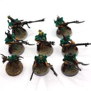 May include: A collection of eight miniature warrior figurines painted in shades of green, gold, and black. Each figure stands on a circular, textured base and is equipped with a long, dark-coloured weapon, with some figures holding blades.