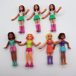 Op de afbeelding: Een verzameling kleine plastic poppen met verschillende outfits. De poppen hebben verschillende huidtinten en haarkleuren. Sommige poppen dragen rokken en laarzen, terwijl anderen shorts en schoenen dragen. De poppen staan in verschillende poses met hun armen uitgestrekt.