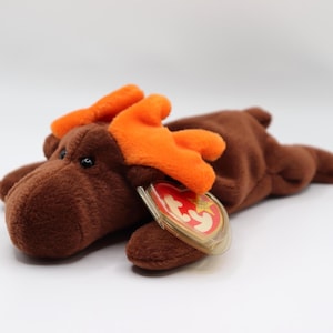 May include: A brown plush moose toy with orange antlers and small black eyes. The moose has a tag attached with red and gold colours. The toy is lying down on a white surface.