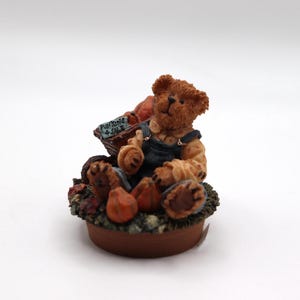 May include: A brown teddy bear figurine sitting on a bed of autumn foliage, holding a basket of pumpkins with a sign that reads "Pumpkins 4 Sale".