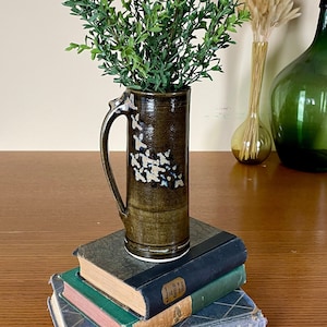 May include: A brown ceramic vase with a white glaze and a handle, filled with green leafy branches. The vase is sitting on a stack of three books with green, red, and blue covers. The books are on a wooden table.