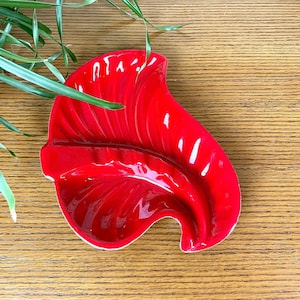 May include: Red ceramic ashtray shaped like a leaf. The ashtray has a white rim and a glossy finish.