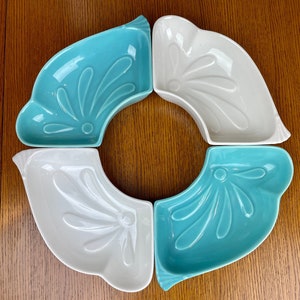 May include: Four ceramic dishes arranged in a circle, two in a light turquoise and two in white. Each dish has a curved shape with a stylized floral design. The dishes are on a wooden surface.