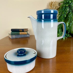May include: A white ceramic coffee pot with a blue and green glaze. The pot has a tall, cylindrical shape with a spout and a handle. There is a matching creamer with a lid.