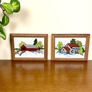 May include: Two framed embroidered artworks. One depicts a red covered bridge with trees and a stream. The other shows a watermill with a red roof, trees, and a water wheel. Both are in wooden frames.