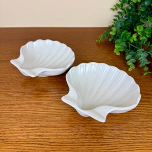 May include: Two white ceramic seashell-shaped dishes. The dishes have a ridged texture and a curved, scalloped edge. The dishes are on a wooden surface, with green foliage in the background. These dishes are ideal for serving small snacks or as decorative pieces.