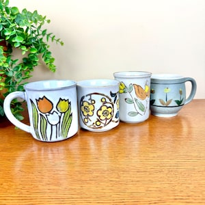 May include: Four ceramic mugs with floral patterns. The mugs feature various colours, including orange, yellow, green, and blue. One mug has a tulip design, while others have abstract floral patterns. The mugs are displayed on a wooden surface.