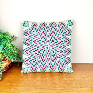 May include: Square decorative pillow with a geometric pattern in shades of green, pink, and white. The pillow is placed on a wooden surface. The pillow's design features a repeating zig-zag pattern.