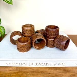 May include: A collection of eight wooden napkin rings. Each ring is a rich brown color and features a carved geometric pattern. The rings are arranged on a white book, creating a rustic and elegant display. The rings are approximately 2 inches in diameter.