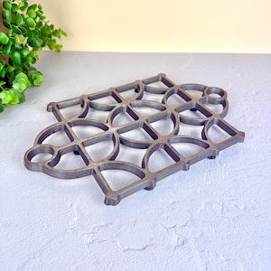 May include: A gray metal trivet with a geometric design of interlocking circles and curved shapes. The trivet is designed to protect surfaces from hot cookware. The trivet measures approximately 20 cm in length and 18 cm in width.
