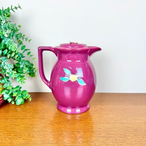 May include: A magenta ceramic jug with a lid and handle. The jug features a floral design with yellow and blue accents. The jug is sitting on a wooden surface.