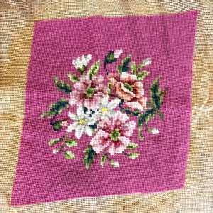 May include: A needlepoint canvas featuring a floral design. The flowers are in shades of pink, white, and peach, with green leaves. The background is a solid magenta color. The canvas is square and has a textured appearance.