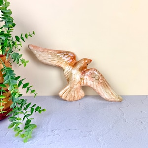 May include: A ceramic dove figurine with outstretched wings, painted in shades of beige, peach, and brown. The bird has detailed feather patterns and a small, red beak. The figurine is a decorative item.