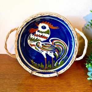 May include: A round, blue ceramic plate with a rooster design in white, green, orange, and brown. The plate has a woven wicker border and handles. The plate is displayed on a metal stand.