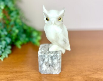 May include: A white stone owl figurine with yellow eyes perched atop a square, grey and white marbled stone base. The owl features carved feather details and a smooth, polished surface, standing upright with its head turned slightly to the side.