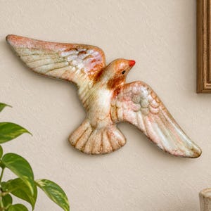 May include: A ceramic wall sculpture of a bird in flight with outstretched wings. The piece features a glossy glaze with blended tones of cream, soft pink, and light brown, accented by a small, bright red beak and a single black eye detail.