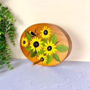 May include: An oval wooden plaque featuring a hand-painted design of bright yellow sunflowers with dark centers and green leaves. A small black and yellow bird is perched among the flowers. A single wooden peg protrudes from the lower center.