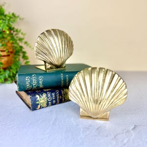 May include: Two gold-colored seashell bookends. The bookends are made of metal and have a detailed, ridged design. One bookend is holding up a dark green book with gold lettering. The other bookend is resting on a light surface.