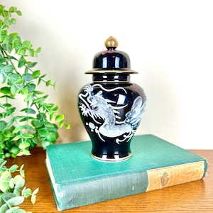 May include: A black ceramic ginger jar with a lid, featuring a white and gray dragon design. The jar has gold accents on the lid and base. It is sitting on a green book.