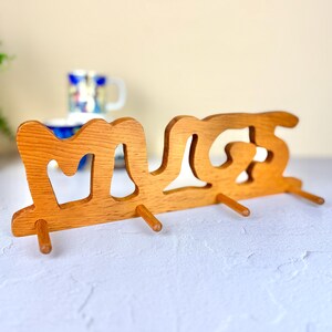 May include: Wooden "Mugs" wall-mounted rack with three pegs for hanging mugs. The rack is made of light brown wood and has the word "Mugs" cut out. A blue and white patterned mug is visible in the background.