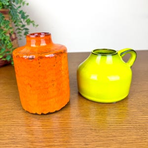 May include: Two ceramic vases. One is a tall, textured orange vase with a narrow neck. The other is a small, rounded, bright yellow vase with a handle. Both vases are on a wooden surface.