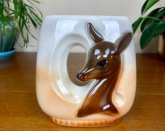 May include: A ceramic vase with a brown deer head design. The vase is white with a brown gradient at the bottom. The deer head is in relief and has a brown glaze.