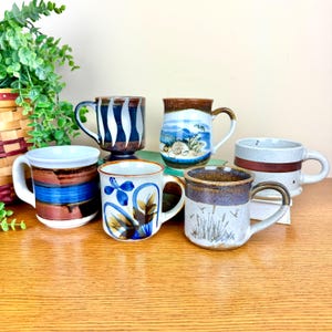 May include: A collection of six ceramic mugs with various designs. The mugs feature colors like blue, brown, white, and beige. Some mugs have striped patterns, while others display floral or landscape artwork. Each mug has a handle for easy gripping.