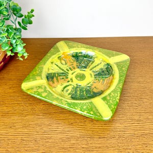 May include: A square, vintage ashtray with a vibrant green and yellow color scheme. The ashtray features a central circular design with raised details in shades of green, yellow, and orange. The ashtray is made of ceramic or glass.