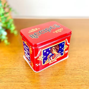 May include: A red rectangular recipe tin with the word "Recipes" on the lid. The tin features a Coca-Cola advertisement with a woman holding an ice cream cone. The tin is decorated with stars and the words "Delicious" and "5¢".