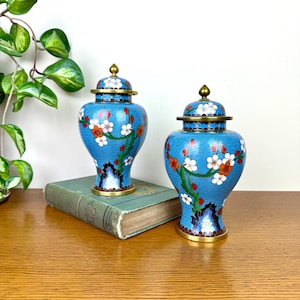 May include: Two blue cloisonné ginger jars with lids. The jars feature a floral design with white and orange blossoms on branches. The jars have gold-colored rims and lids, and are placed on a vintage book.