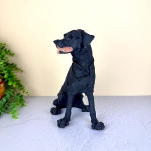 May include: A black dog figurine with a textured surface, depicted in a seated position with its head turned to the side. The sculpture features a visible pink tongue protruding from its mouth and a collar around its neck, rendered in a solid black finish.