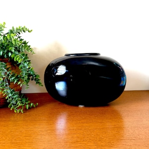 May include: A glossy, black ceramic vase with a rounded, oval shape. The vase has a wide opening at the top and sits on a wooden surface. A green plant in a woven basket is visible on the left side.