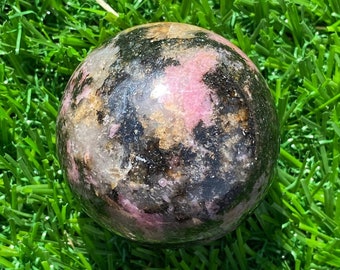 May include: A polished spherical stone featuring a mottled pattern of deep black, soft pink, and translucent white mineral inclusions. The surface of the sphere is smooth and reflective, showcasing natural variations in color and texture throughout.