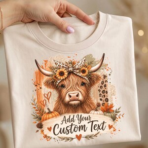 May include: A cream-colored t-shirt featuring a graphic of a Highland cow wearing a leopard print bow and sunflower crown. The design includes pumpkins, autumn leaves, and the text Add Your Custom Text with small orange hearts.
