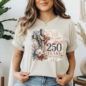 Könnte beinhalten: Ein beiges Kurzarm-T-Shirt mit einer Grafik eines Holzkreuzes, das mit einer amerikanischen Flagge und Blumen geschmückt ist, sowie dem Text: So if the Son sets you free, you will be free indeed. John 8:36. 250 YEARS OF GOD'S FAITHFULNESS 1776-2026.