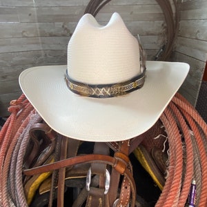 May include: A white cowboy hat with a brown leather band and feather accents sits on top of a pile of brown leather straps and red rope.
